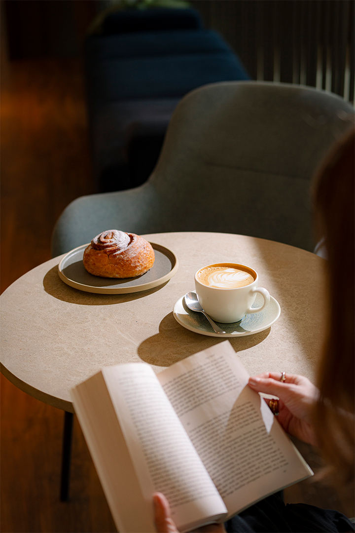 A woman reading a book with a coffee and a pastry on a table in front of her