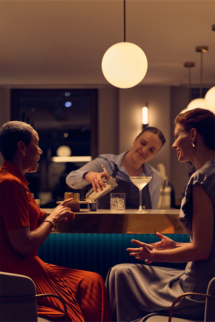 Two women sat at a bar smiling, with cocktails on the bar next to them