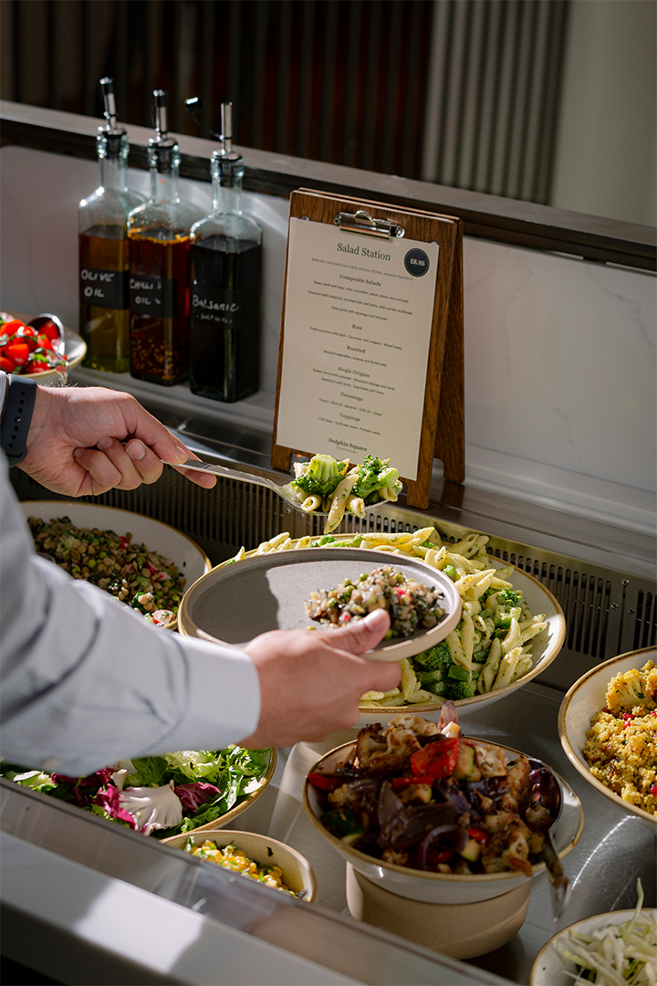 A person spooning salad onto a plate from the Dolphin House Cafe & Bar Salad Station